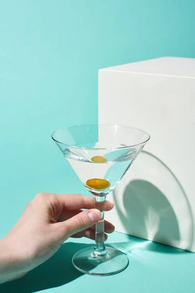 Vue partielle de la femme tenant verre transparent avec cocktail et olive près du cube blanc sur fond turquoise — Photo de stock