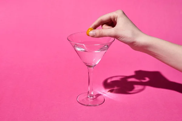 Cropped view of woman putting olive in glass with cocktail on pink background — Stock Photo