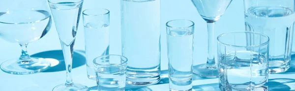 Panoramic shot of transparent glasses with clear water on blue background — Stock Photo