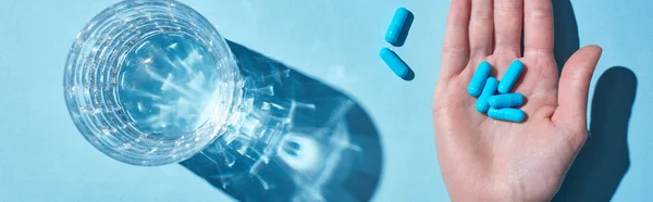 Cropped view of woman holding blue pills on palm near glass with water on blue background, panoramic shot — Stock Photo