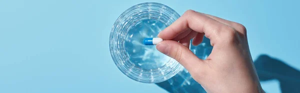 Vista recortada de la mujer poner píldora en vidrio con agua sobre fondo azul, tiro panorámico - foto de stock
