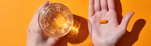 Cropped view of woman holding pill on palm near glass with water on orange background, panoramic shot — Stock Photo