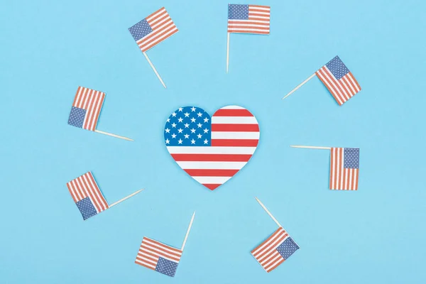 Marco redondo de papel cortado banderas americanas decorativas en palos de madera y corazón hecho de estrellas y rayas sobre fondo azul - foto de stock