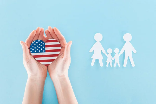 Vista ritagliata della donna che tiene il cuore fatto di bandiera nazionale americana vicino alla famiglia taglio carta bianca su sfondo blu — Foto stock