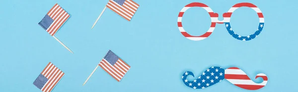 Colpo panoramico di occhiali e baffi fatti di stelle e strisce vicino a bandiere decorative americane su bastoni di legno su sfondo blu — Foto stock