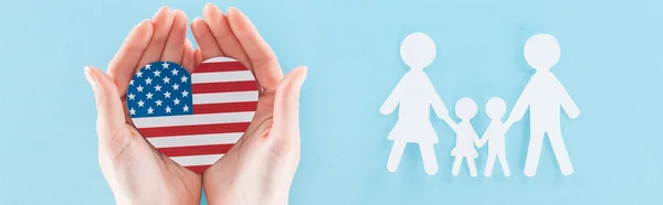 Plano panorámico de la mujer sosteniendo el corazón hecho de bandera nacional americana cerca de la familia de corte de papel blanco sobre fondo azul - foto de stock