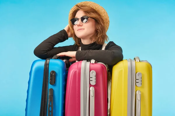 Fille rêveuse dans des lunettes de soleil et chapeau de paille près de sacs de voyage multicolores isolés sur bleu — Photo de stock