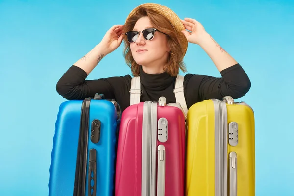 Fröhliches Mädchen mit Sonnenbrille und Strohhut in der Nähe bunter Reisetaschen auf blauem Grund — Stockfoto