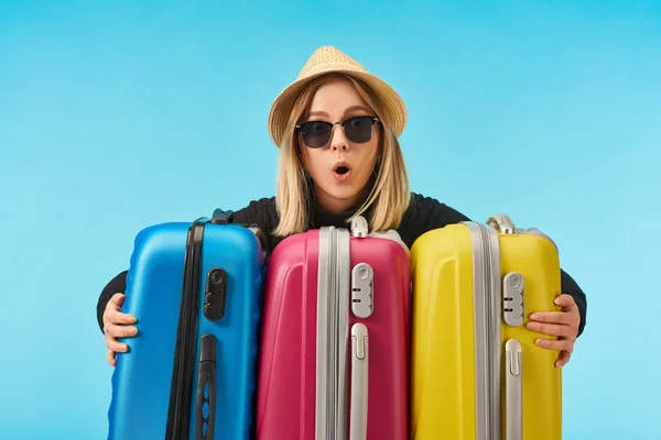 Menina chocada em óculos de sol e chapéu de palha perto de sacos de viagem multicoloridos isolados em azul — Fotografia de Stock
