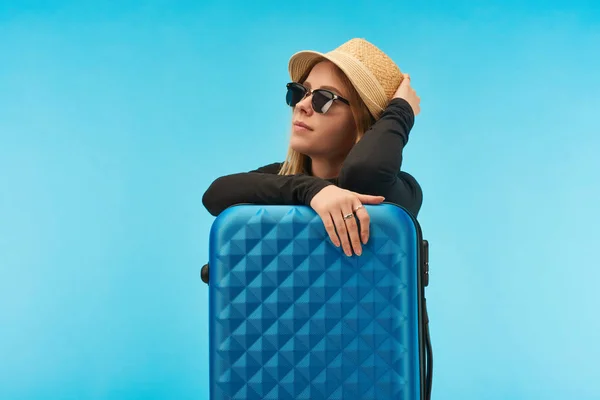 Блондинка в сонцезахисних окулярах і солом'яному капелюсі біля синьої валізи ізольована на синьому — стокове фото