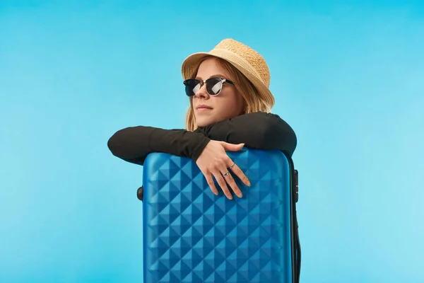 Chica rubia soñadora en gafas de sol y sombrero de paja cerca de la bolsa de viaje azul aislado en azul - foto de stock