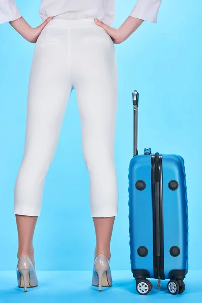 Вид ззаду жінки, що стоїть з руками на стегнах біля синього барвистого дорожнього сумки на синьому фоні — стокове фото