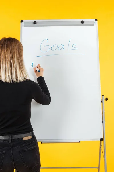 Back view of businesswoman standing near white flipchart and writing goals list — Stock Photo
