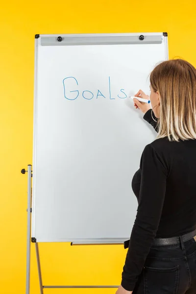 Vista posterior de la mujer de negocios de pie cerca del rotafolio blanco, escribiendo objetivos palabra - foto de stock