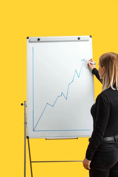 Rückseite einer Geschäftsfrau, die neben weißem Flipchart steht, Wachstumsgrafik zeichnet, isoliert auf gelb steht — Stockfoto