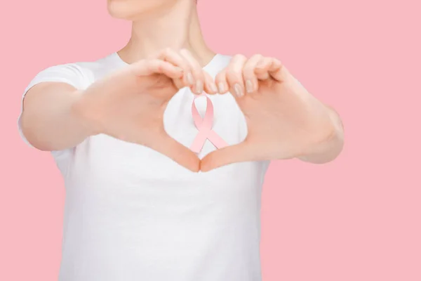 Partial view of woman in white t-shirt doing heart sign with hands around pink breast cancer ribbon isolated on pink — Stock Photo