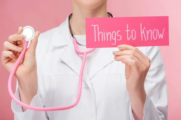 Vista parziale del medico in mantello bianco e stetoscopio in possesso di carta con cose da sapere lettering isolato su rosa — Foto stock