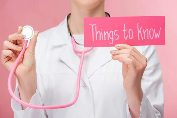 Partial view of doctor in white coat and stethoscope holding card with things to know lettering isolated on pink — Stock Photo