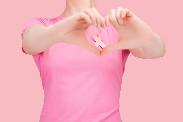 Обрезанный вид женщины в розовой футболке, показывающей знак сердца с руками вокруг ленты рака груди, изолированной на розовый — стоковое фото