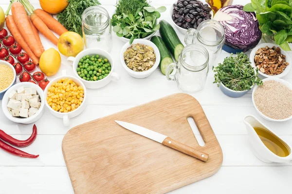 Vista dall'alto di frutta e verdura fresca vicino a vasi di vetro su tavolo bianco in legno con tagliere e coltello — Stock Photo