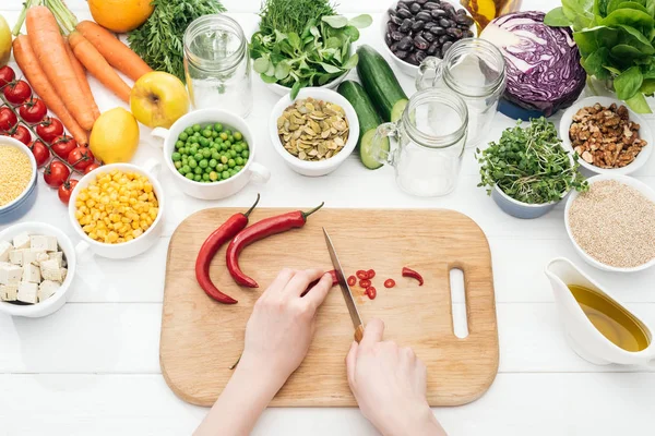 Abgeschnittene Ansicht einer Frau, die Chilischoten auf einem hölzernen Schneidebrett auf weißem Tisch schneidet — Stockfoto