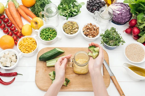 Cropped view of woman adding chopped cucumber to couscous in glass jar on wooden chopping board — Stock Photo