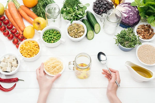 Cropped view of woman holding bowl with couscous near glass jar on wooden white table — Stock Photo