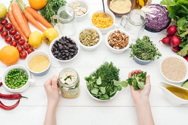 Vue recadrée de femme ajoutant des herbes dans un bocal avec salade sur table blanche en bois — Photo de stock