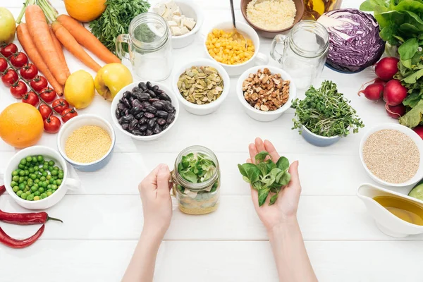 Vista ritagliata di donna che aggiunge erbe in vaso di vetro con insalata su tavolo bianco in legno — Foto stock