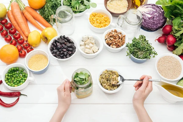 Cropped view of woman adding pumpkin seeds in jar with salad on wooden white table — Stock Photo