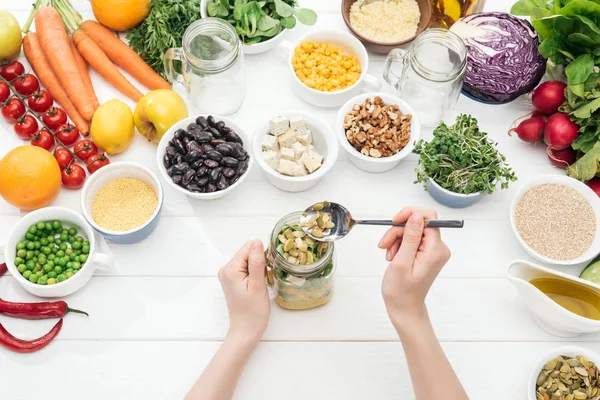 Cropped view of woman adding pumpkin seeds in glass jar with salad on wooden white table — Stock Photo