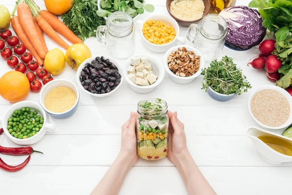 Vista recortada de la mujer en la celebración de frasco de vidrio con ensalada en la mesa blanca de madera — Stock Photo