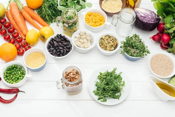 Vista superior de frutas y verduras cerca de frasco de vidrio con nueces y plato con rúcula verde sobre mesa blanca de madera - foto de stock