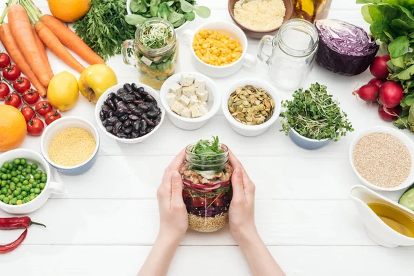 Vista cortada da mulher segurando frasco de vidro com salada fresca na mesa branca de madeira — Fotografia de Stock