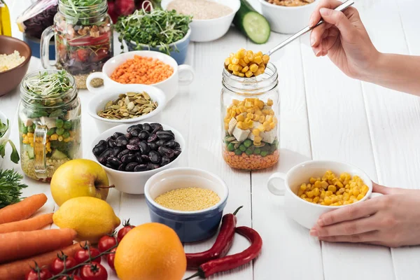 Vista ritagliata di donna che aggiunge mais in vaso di vetro con insalata su tavolo bianco in legno — Foto stock