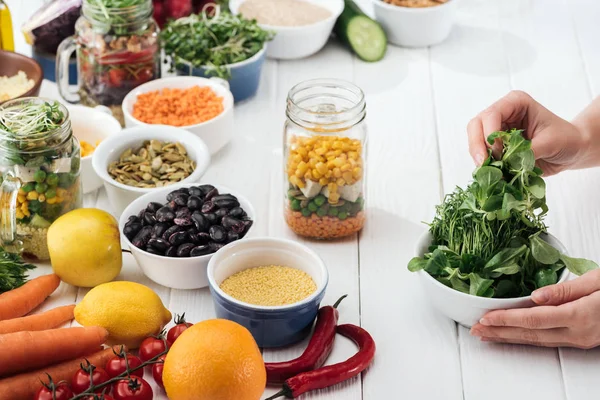 Cropped view of woman taking greenery from bowl on wooden white table — Stock Photo