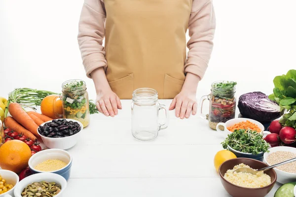 Cropped view of woman in apron with empty jar and vegetables on wooden table isolated on white — Stock Photo