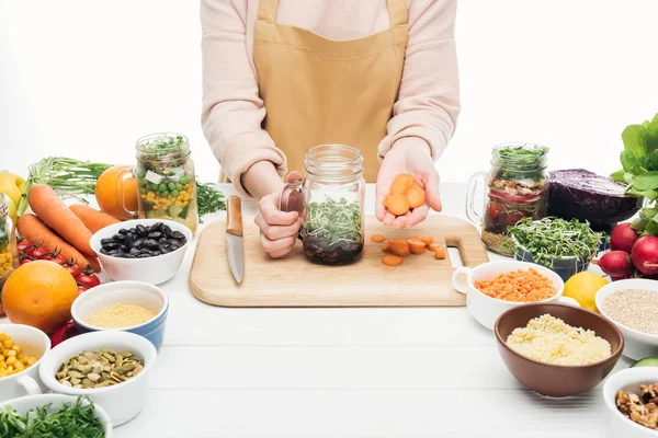 Vista ritagliata di donna in grembiule aggiungendo carota tagliata a fette in vaso con insalata su tavolo di legno isolato su bianco — Foto stock