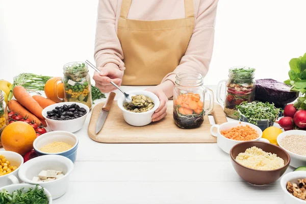 Cropped view of woman in apron adding pumpkin seeds in jar on wooden table isolated on white — Stock Photo