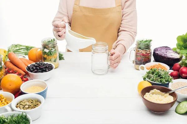 Vista recortada de la mujer en delantal añadiendo aceite en vidrio vacío sobre mesa de madera aislada en blanco — Stock Photo