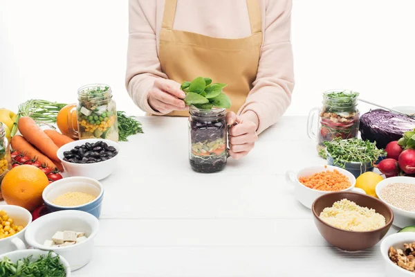 Abgeschnittene Ansicht einer Frau in Schürze, die grüne Blätter in Glasgefäßen auf einem Holztisch hinzufügt, isoliert auf weiß — Stockfoto