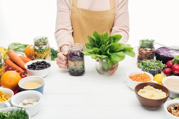 Cropped view of woman in apron holding glass jar with salad and green leaves on wooden table isolated on white — Stock Photo