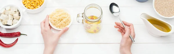 Cropped view of woman holding bowl with couscous near glass jar on wooden white table, panoramic shot — Stock Photo