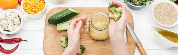 Cropped view of woman adding chopped cucumber to couscous in glass jar on wooden chopping board, panoramic shot — Stock Photo