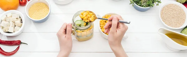 Vue recadrée de femme ajoutant du maïs dans un bocal avec salade sur table blanche en bois, vue panoramique — Photo de stock