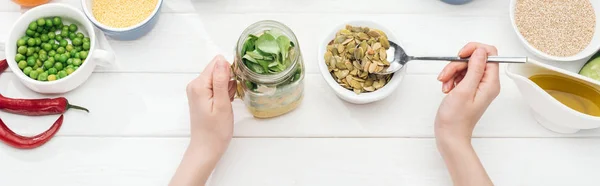 Vue recadrée de femme ajoutant des graines de citrouille dans un bocal avec salade sur une table blanche en bois, vue panoramique — Photo de stock