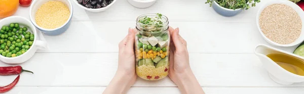 Cropped view of woman holding glass jar with fresh salad on wooden white table, panoramic shot — Stock Photo