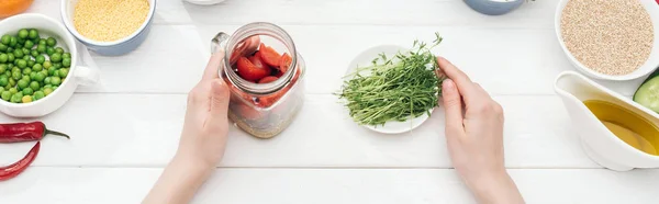 Vista recortada de la mujer sosteniendo frasco con ensalada y brotes verdes en la mesa blanca de madera, tiro panorámico — Stock Photo