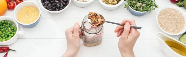 Cropped view of woman adding nuts in glass jar with salad on wooden white table, panoramic shot — Stock Photo