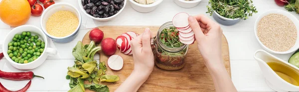 Cropped view of woman adding radish slices in jar with salad on wooden white table, panoramic shot — Stock Photo